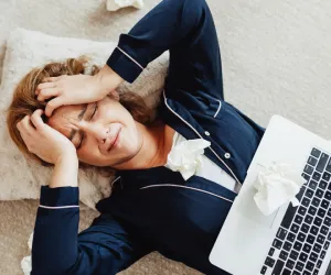 Femmeallongeeausolenpyjamalesmainssurlateteavecunordinateurportableetdesmouchoirsexprimantlafatiguechroniqueetlepuisementemotionnelliesauburnout.Nadege