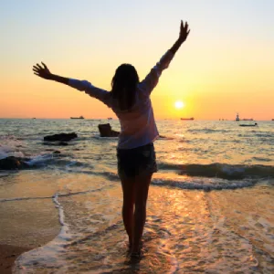 Femme debout au bord de la mer, levant les bras vers le ciel au coucher du soleil, symbole de libération émotionnelle
