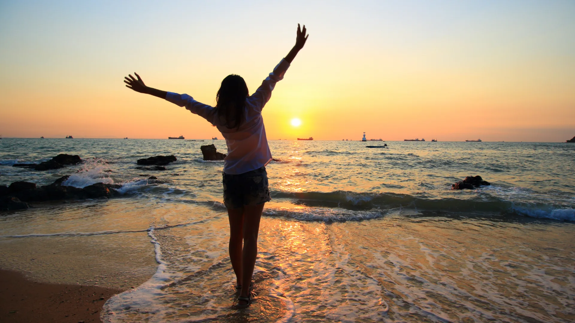 Femme debout au bord de la mer, levant les bras vers le ciel au coucher du soleil, symbole de libération émotionnelle