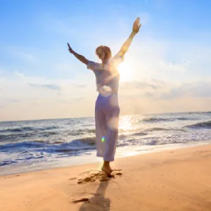 Femme levant les bras au soleil couchant sur une plage, symbole de libération émotionnelle et de bienêtre grâce au ThetaHealing
