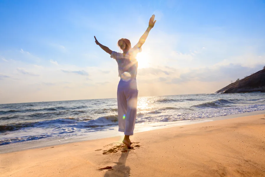 Femme levant les bras au soleil couchant sur une plage, symbole de libération émotionnelle et de bienêtre grâce au ThetaHealing