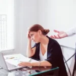 Femme d’affaires fatiguée devant son ordinateur, la tête dans la main, écoutant un collègue lui parler avec insistance, symbole de surcharge mentale et d’épuisement professionnel.
