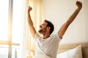 Homme souriant et énergique s’étirant au réveil dans sa chambre, symbole de repos réparateur, équilibre de vie et regain d’énergie.